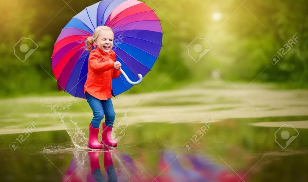 Criança feliz engraçado ba por menina com um guarda-chuva multicolorido pulando em poças de borracha botas e rindo
