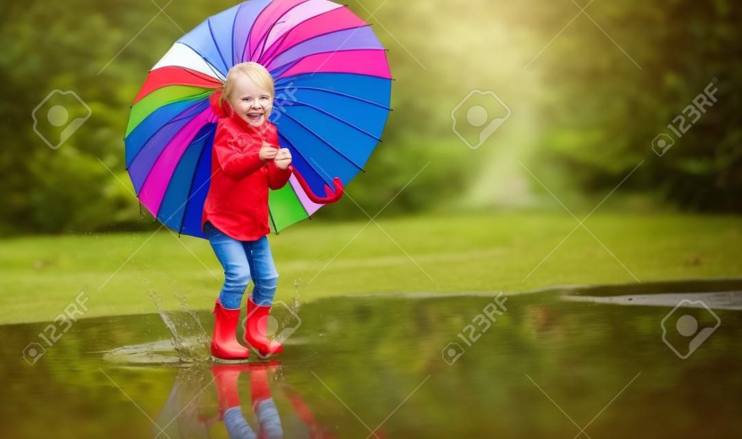Happy funny ba child by girl with a multicolored umbrella jumping on puddles in rubber boots and laughing