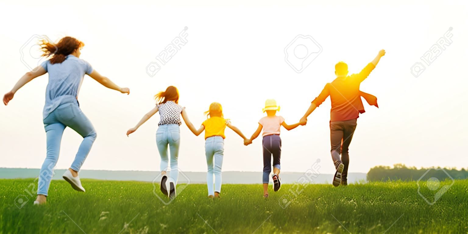 Padres felices irreconocibles con hijas en ropa casual corriendo hacia atrás tomados de la mano mientras disfrutan de tiempo juntos en el prado verde en las noches de verano