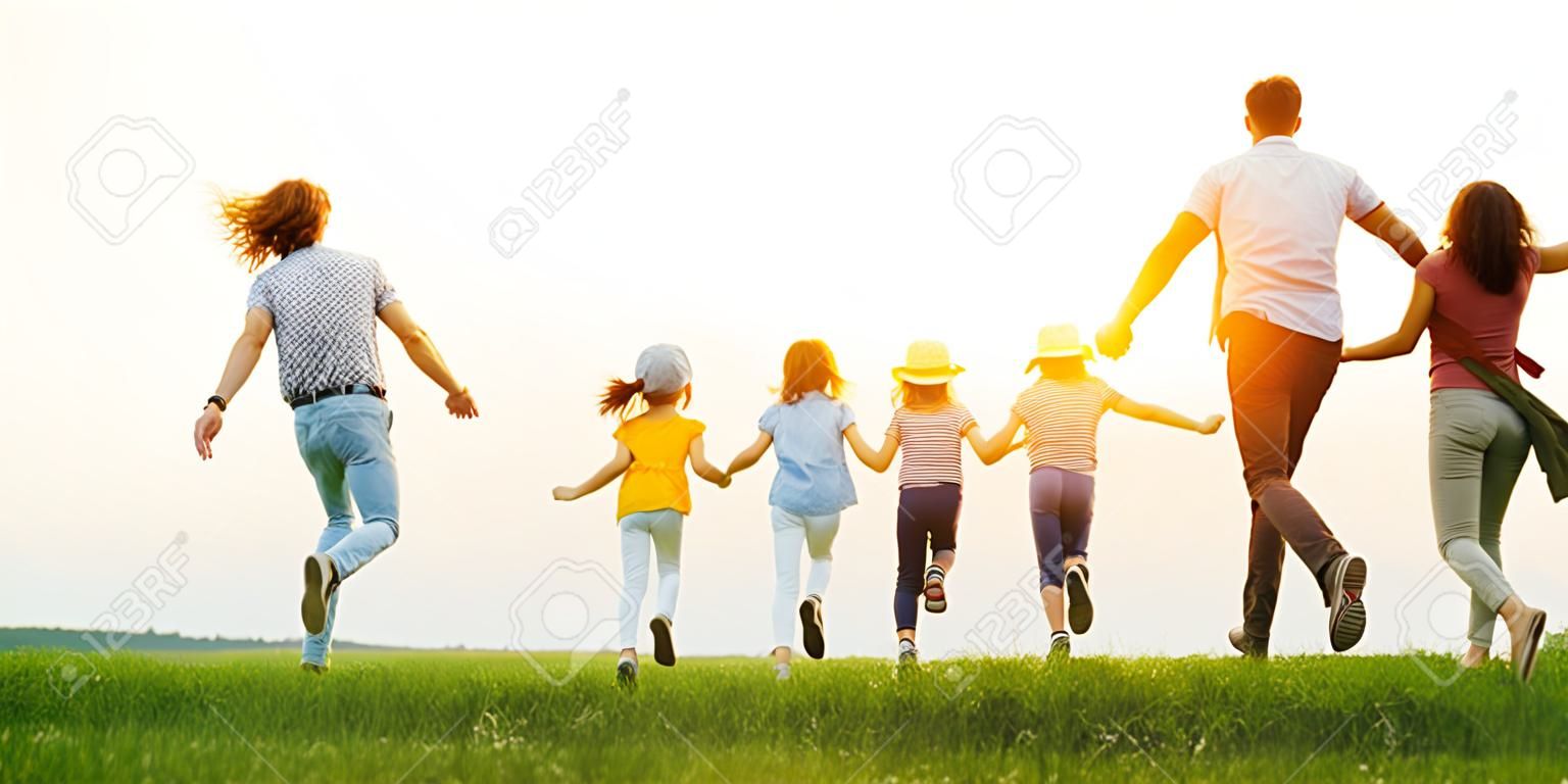 Padres felices irreconocibles con hijas en ropa casual corriendo hacia atrás tomados de la mano mientras disfrutan de tiempo juntos en el prado verde en las noches de verano