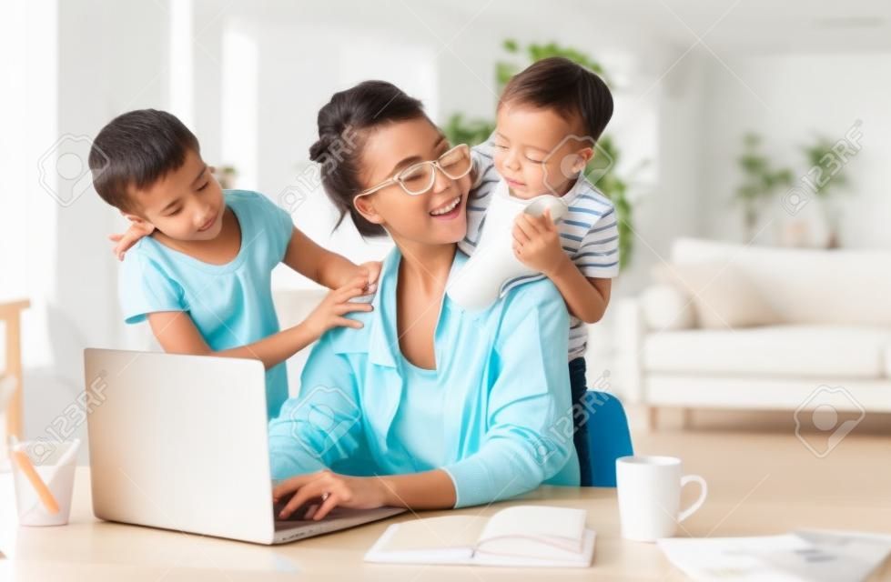 Menina perturbada abraçando e pequeno filho mostrando brinquedo para mãe ocupada freelancer sentado na mesa com xícara de café e usando laptop contra interior turvo de luz apartamento moderno