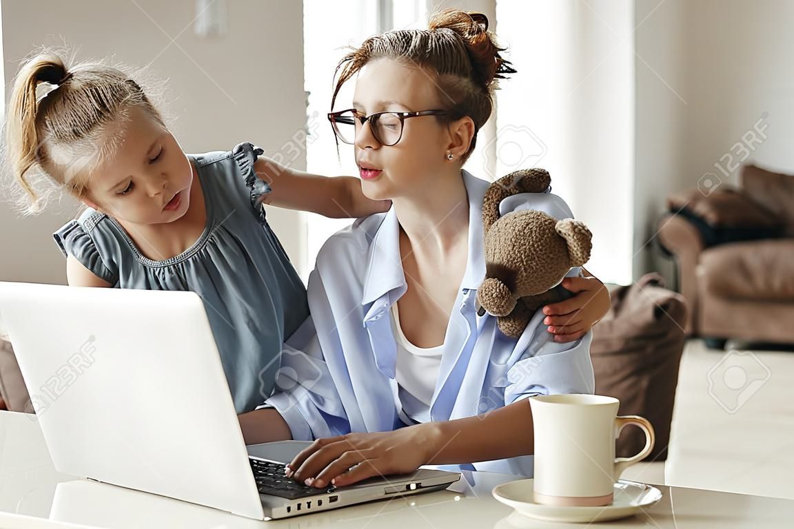 Niña molesta abrazos e hijo pequeño que muestra el juguete a la ocupada madre independiente sentada en la mesa con una taza de café y usando la computadora portátil contra el interior borroso del apartamento moderno y ligero