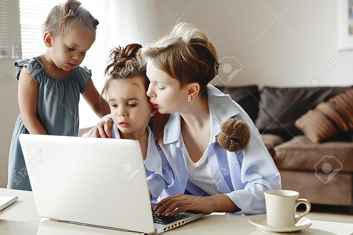 Upset girl hugging and small son showing toy to busy mother freelancer sitting at table with cup of coffee and using laptop against blurred interior of light modern apartment