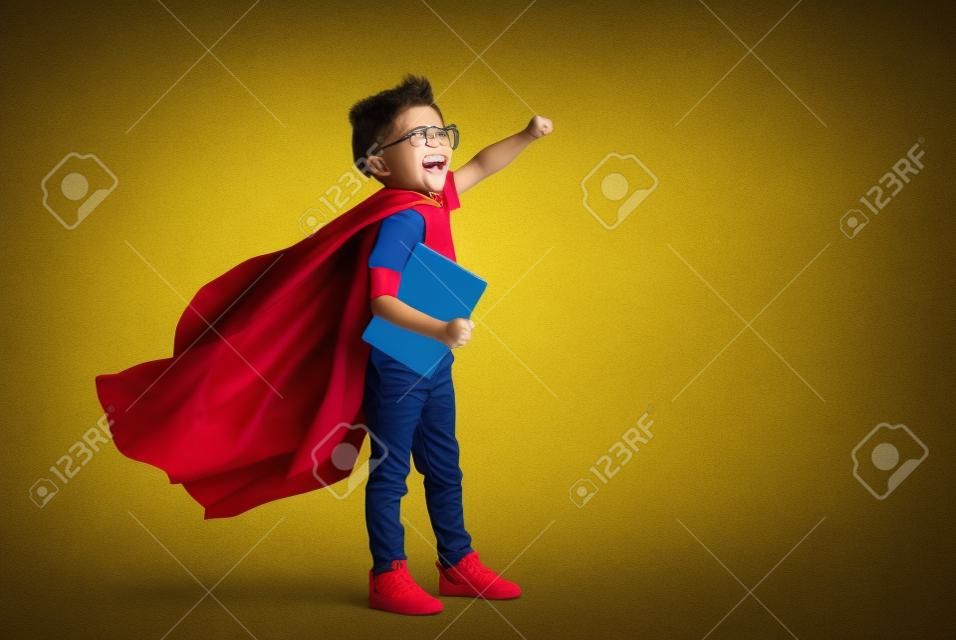 黄色の背景に学校の研究の準備ができている間、笑顔で拳を上げるスーパーヒーローマントの全身少年