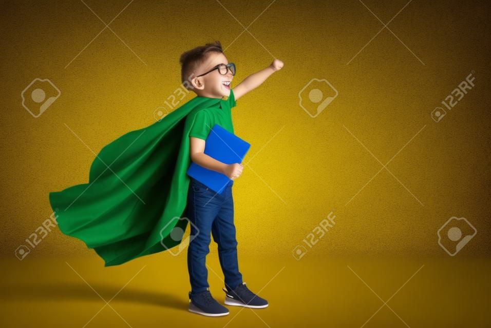 黄色の背景に学校の研究の準備ができている間、笑顔で拳を上げるスーパーヒーローマントの全身少年