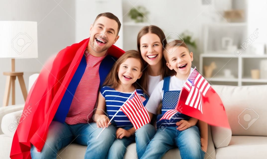 Jeunes parents de famille américains heureux et deux petits enfants assis sur un canapé à la maison avec des drapeaux des états-unis et souriant à la caméra tout en célébrant le jour de l'indépendance. Concept de vacances américain patriotique