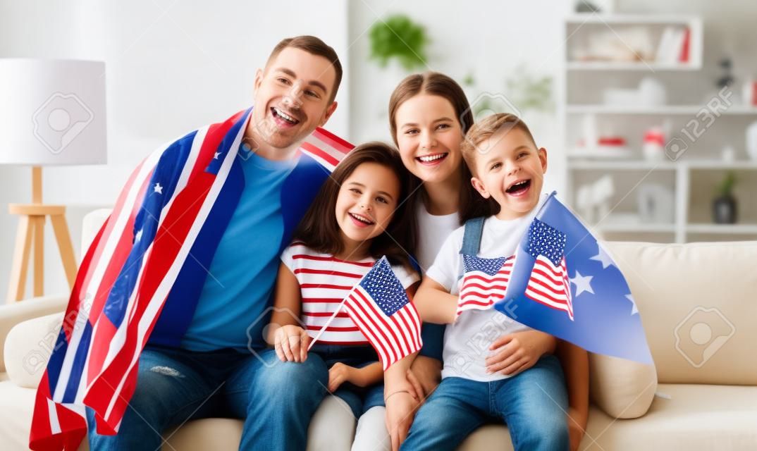 Jóvenes padres de familia estadounidenses felices y dos niños pequeños sentados en un sofá en casa con banderas de estados unidos y sonriendo a la cámara mientras celebran el Día de la Independencia. Concepto patriótico de vacaciones en EE. UU.