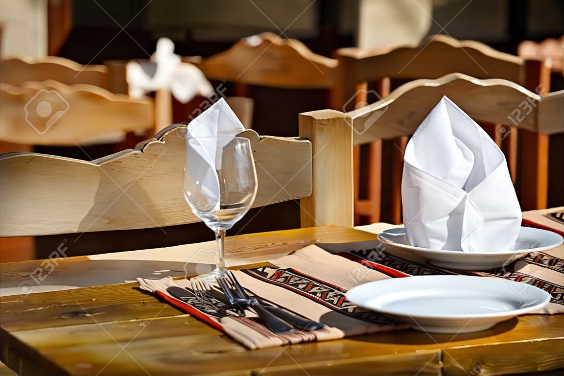 Two empty white plates, two glasses with napkins standing on a wooden table. Street restaurant.