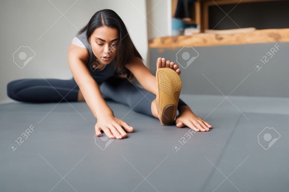 Close up of young woman practicing yoga, sitting in Head to Knee Forward Bend exercise, Janu Sirsasana pose, working out, wearing sportswear, grey pants, bra, indoor, home interior background