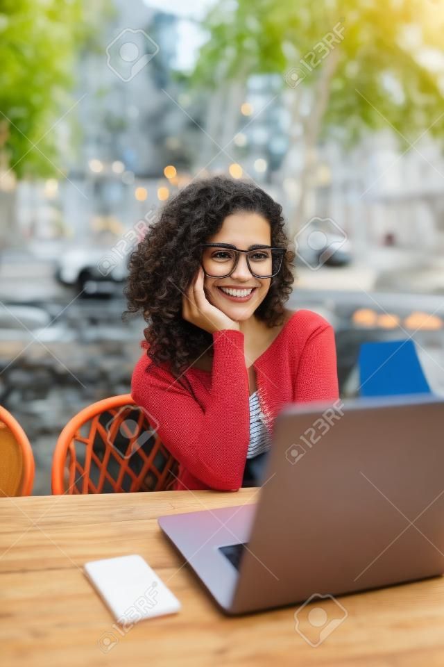 Portrait of latin woman in glasses working with her laptop outdoors. Charming freelancer woman with curly hair is sitting in street bar and doing her remote work on the netbook