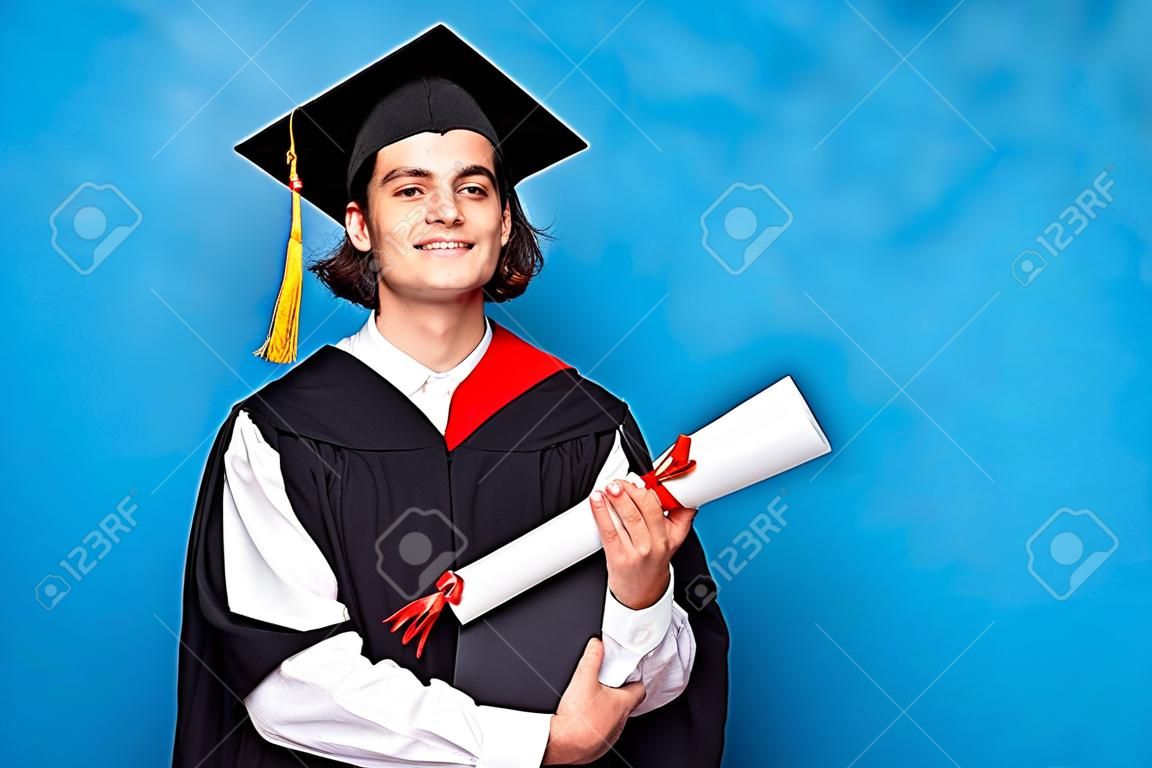 Afgestudeerde mannelijke student draagt zwarte mantel en mortierboard in blauw shirt met zijn blauwe diploma. Plaats voor reclame