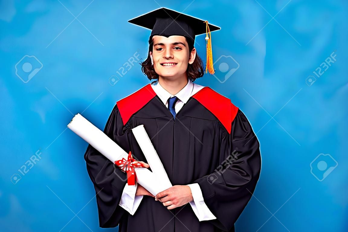 Afgestudeerde mannelijke student draagt zwarte mantel en mortierboard in blauw shirt met zijn blauwe diploma. Plaats voor reclame