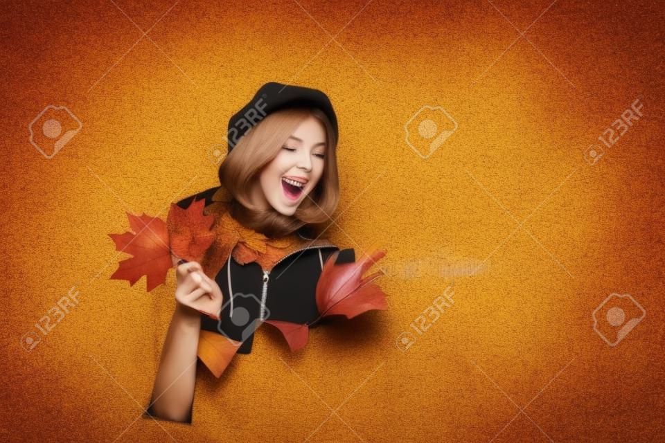 Beleza e moda. Menina de outono feliz pisca. Mulher alegre mostra o sinal ok. Menina com folhas olhando através do buraco de papel. Menina piscando. Hora do outono.