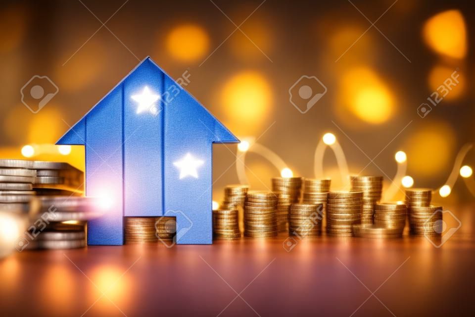 real estate price increase or mortgage interest rates going up conceptual image, cardboard house in front of stacks of coins with fairy lights in the background