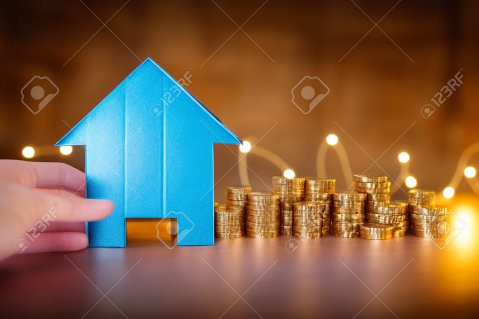 real estate price increase or mortgage interest rates going up conceptual image, cardboard house in front of stacks of coins with fairy lights in the background