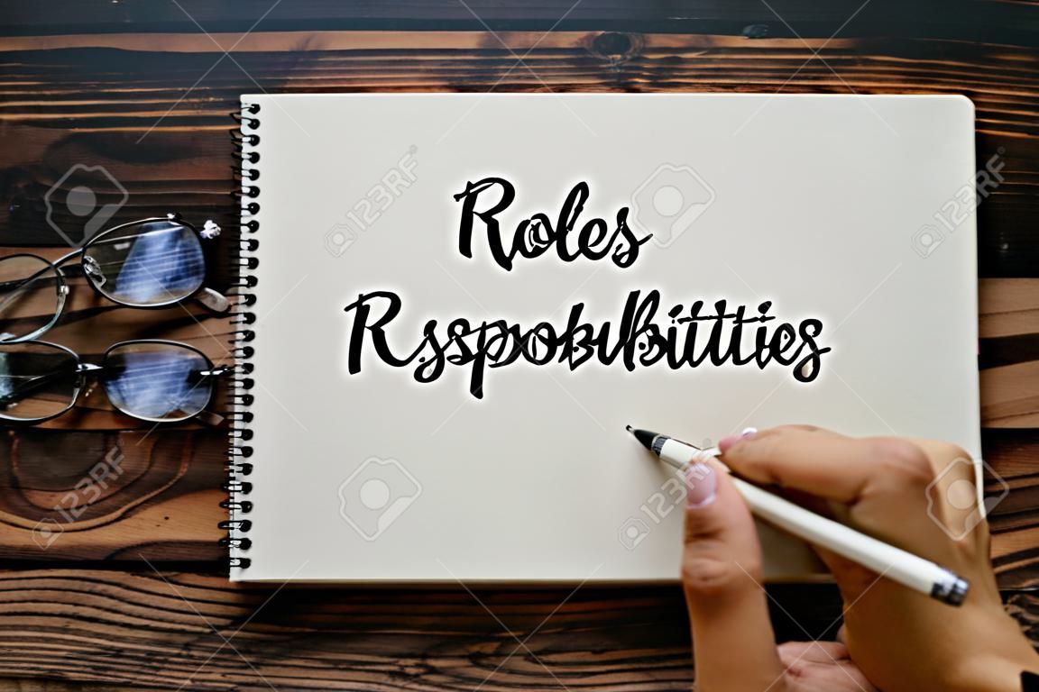 Top view of glasses and hand holding pen writing Roles and Responsibilities on notebook on wooden background.