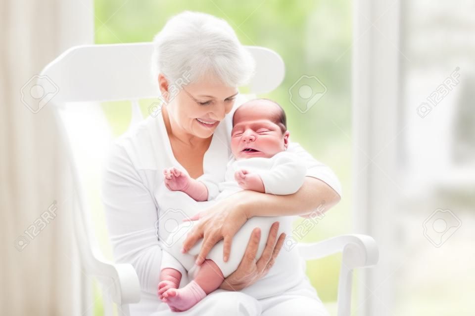 Liebevolle Großmutter sang ein Lied, um ihr neugeborenes Baby Enkel sitzen in einem weißen Schaukelstuhl neben einem großen Garten Fenster