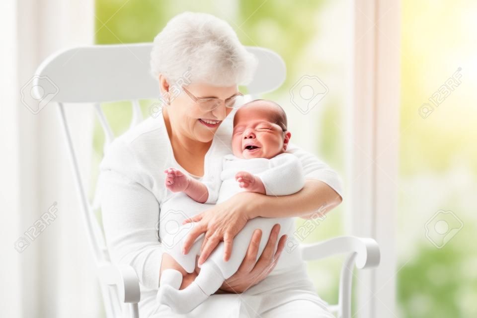 Liebevolle Großmutter sang ein Lied, um ihr neugeborenes Baby Enkel sitzen in einem weißen Schaukelstuhl neben einem großen Garten Fenster
