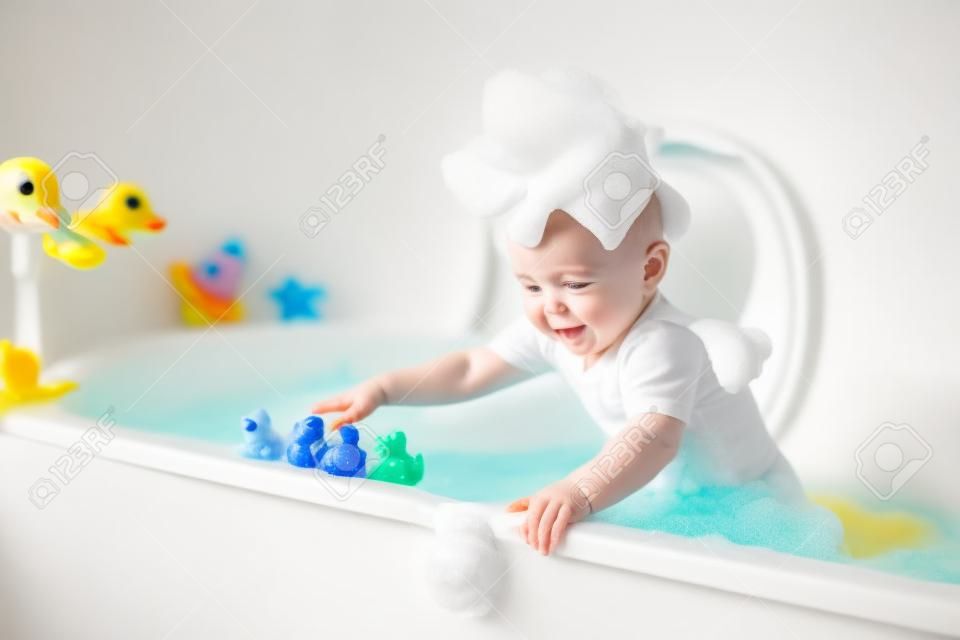 Schattig kleine baby jongen nemen bad spelen met schuim en kleurrijke rubberen eend speelgoed in een witte zonnige badkamer