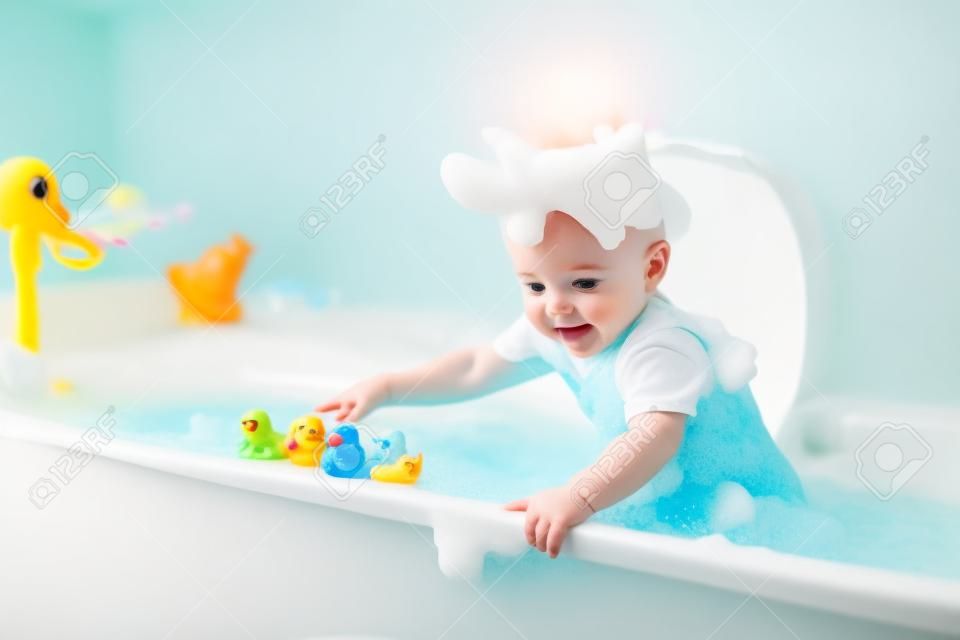 Piccolo neonato sveglio che cattura bagno giocare con i giocattoli schiuma e anatra di gomma colorata in un bagno di sole bianco