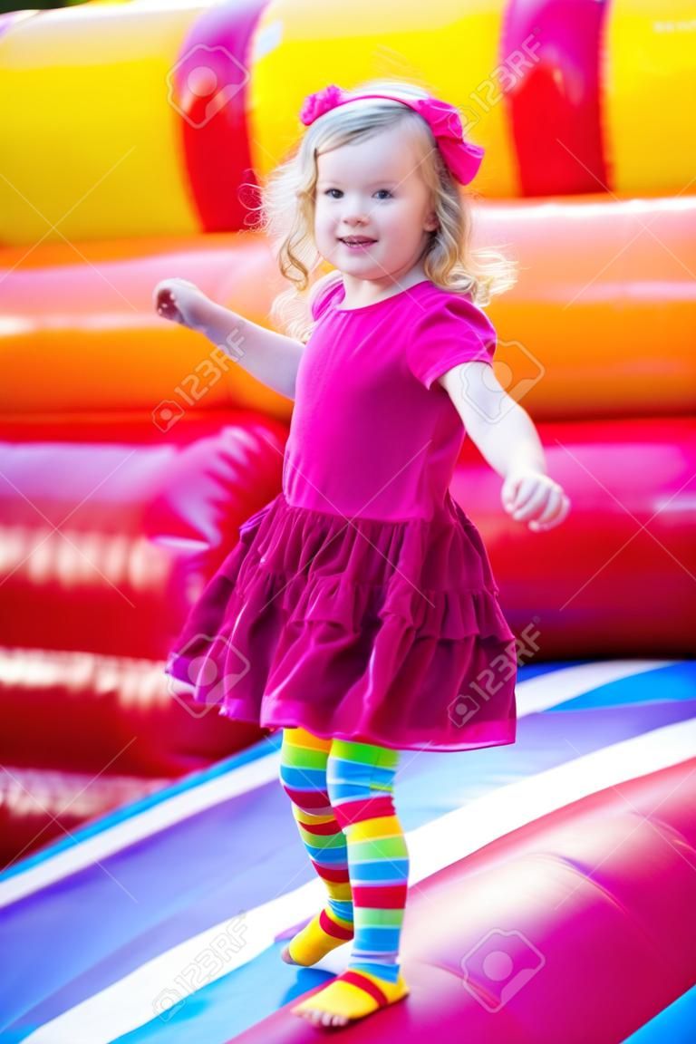 Niña preescolar divertido lindo en un vestido de colores de juego, saltando y saltando en un castillo inflable divirtiéndose en una fiesta de cumpleaños de los niños en un parque infantil en verano
