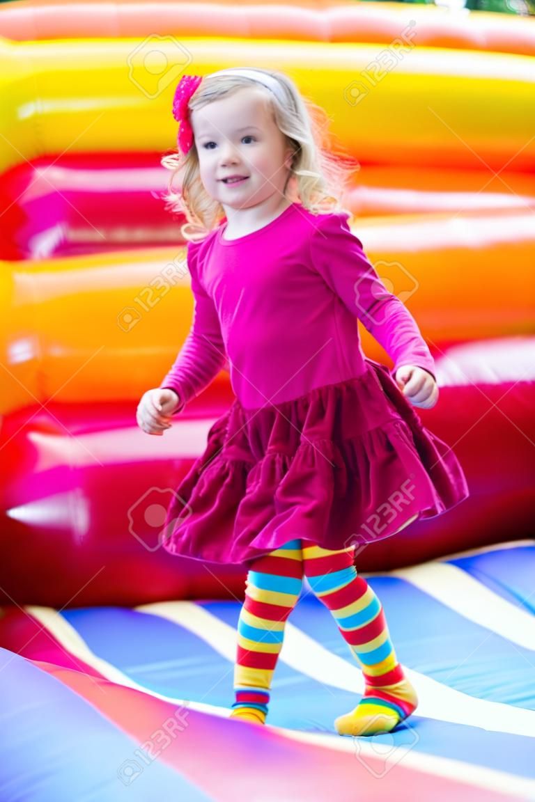 Menina pré-escolar engraçado bonito em um vestido colorido jogando, saltando e saltando em um castelo inflável se divertindo em uma festa de aniversário de crianças em um parque infantil no verão