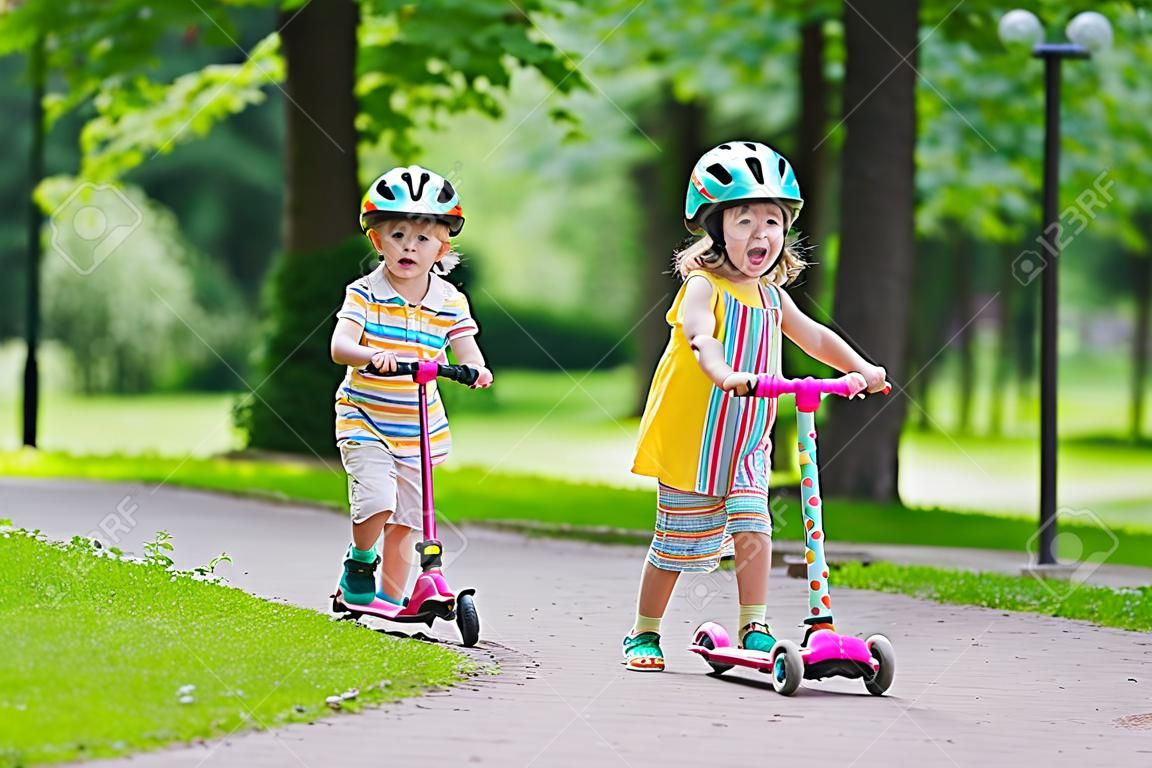 As crianças aprendem a andar de scooter em um parque no dia de verão ensolarado. Menino pré-escolar e menina em capacete de segurança montando um rolo. As crianças brincam ao ar livre com scooters. lazer ativo e esporte ao ar livre para criança.