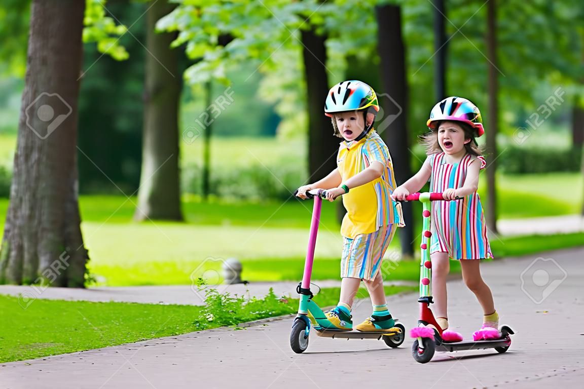 Дети учатся ездить скутер в парке на солнечный летний день. Дошкольника мальчик и девочка в каске верхом ролик. Дети играют на открытом воздухе с скутеров. Активный отдых и спорт на открытом воздухе для ребенка.