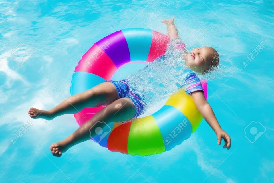 Happy little girl playing with colorful inflatable ring in outdoor swimming pool on hot summer day. Kids learn to swim. Children wearing sun protection rash guard relaxing in tropical resort