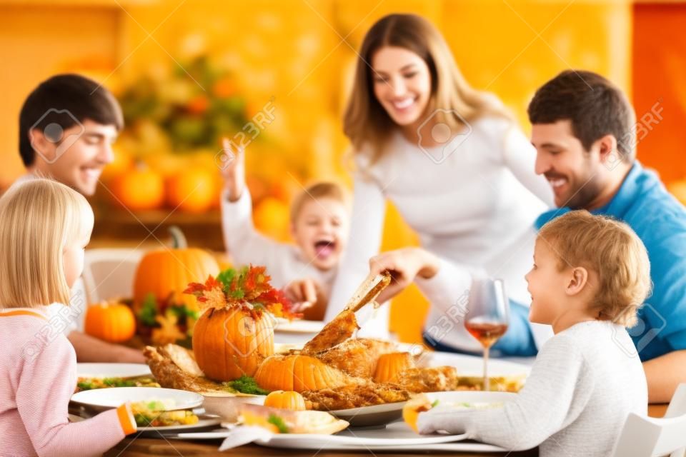 Familie mit Kindern, die Thanksgiving-Dinner essen. Gebratener Truthahn und Kürbiskuchen auf Esstisch mit Herbstdekoration. Eltern und Kinder beim festlichen Essen. Vater und Mutter schneiden Fleisch.