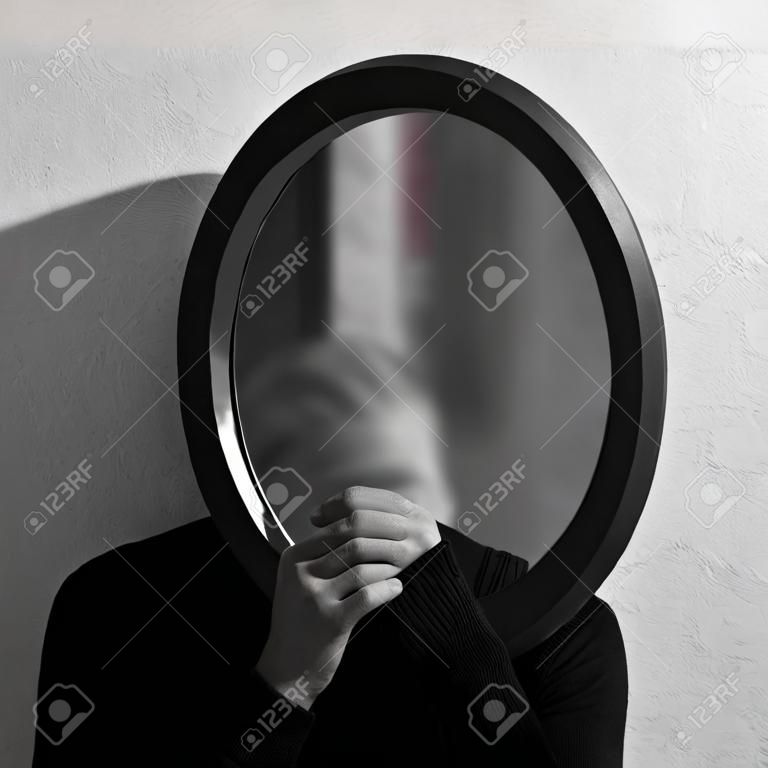 Schwarz-Weiß-Studioporträt eines jungen Mannes, der einen ovalen Spiegel im Gesicht hält. Hintergrund der strukturierten Wand.