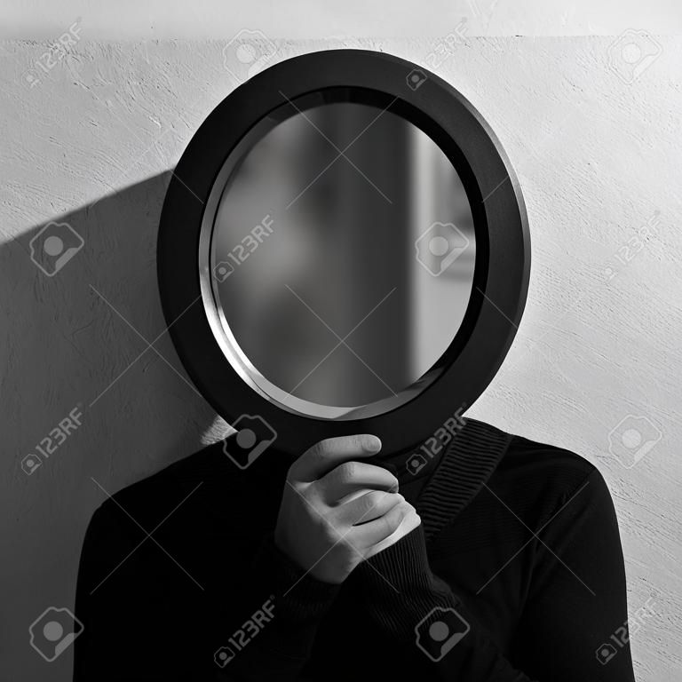 Schwarz-Weiß-Studioporträt eines jungen Mannes, der einen ovalen Spiegel im Gesicht hält. Hintergrund der strukturierten Wand.