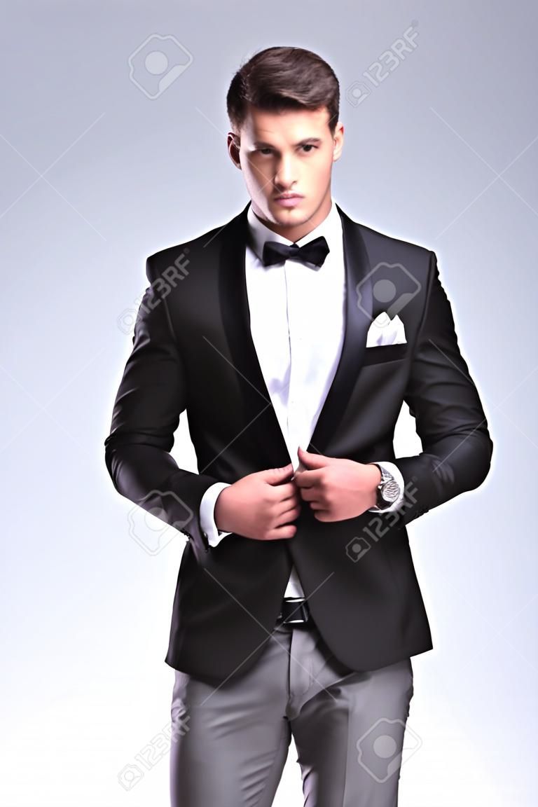 あなたを見ている間彼のジャケットをボタンのタキシードでエレガントなファッションの若い男。灰色の背景に