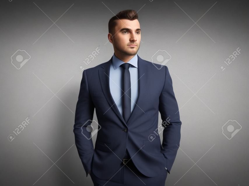 portrait of relaxed businessman in navy suit looking to side while standing on grey wallpaper background with hands in pockets