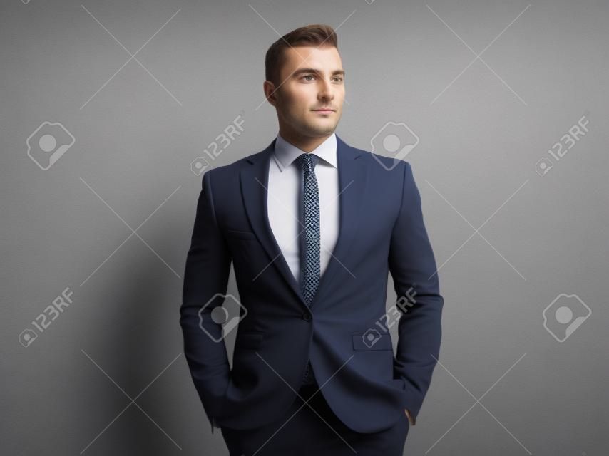 portrait of relaxed businessman in navy suit looking to side while standing on grey wallpaper background with hands in pockets