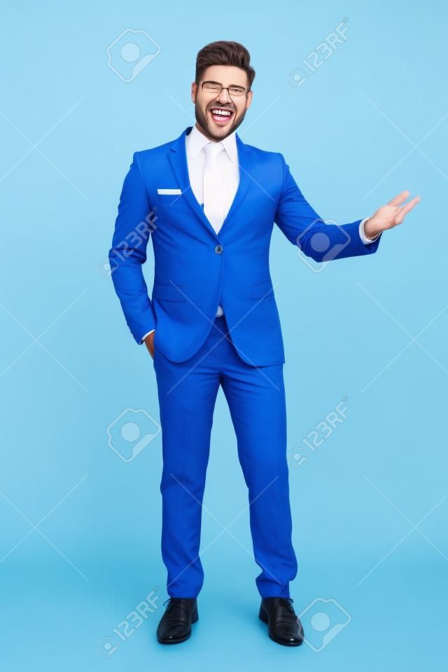 Cheerful businessman presenting with his hand in his pocket while laughing and wearing a blue suit, standing on white studio background