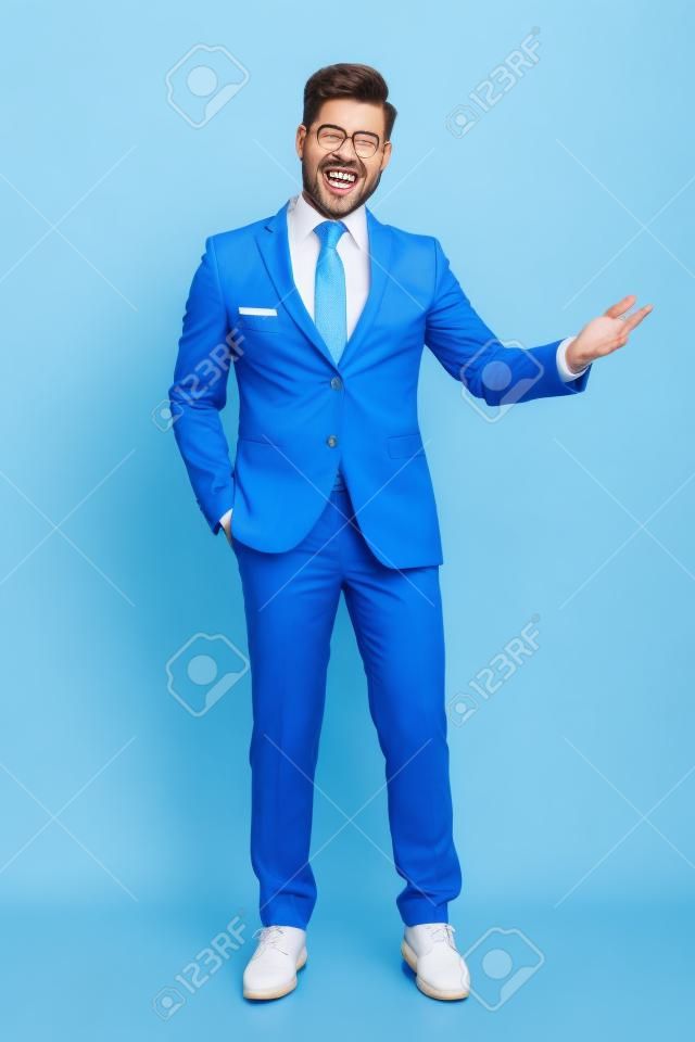Empresário alegre apresentando com a mão no bolso enquanto ri e vestindo um terno azul, de pé no fundo branco do estúdio
