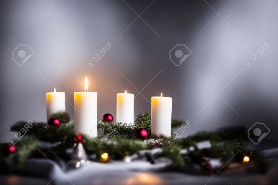 Una delle quattro candele sta bruciando per il primo Avvento su rami di abete con decorazioni natalizie su sfondo grigio, spazio per la copia, messa a fuoco selezionata, profondità di campo ridotta