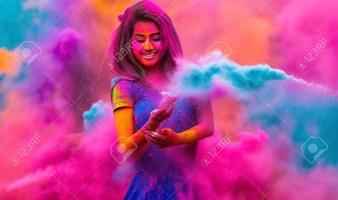 Beautiful young woman celebrating Holi paint fest with colorful powder covering her face and a cloud of dust. On black with rainbow bursts of color