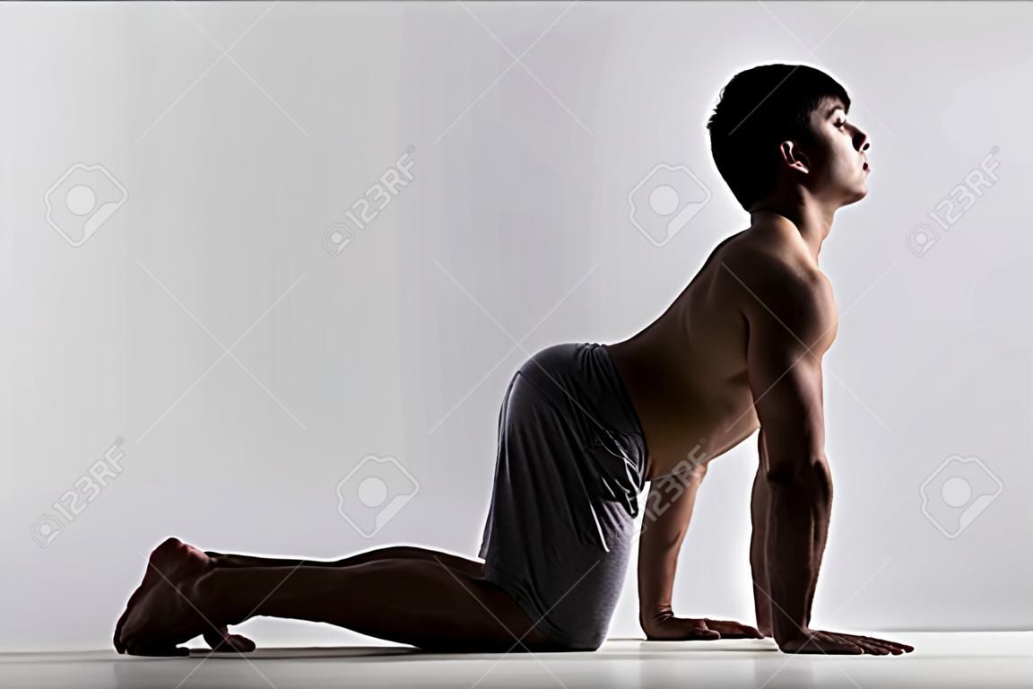 Sporty muscular young man working out, yoga, pilates, fitness training, bend in Cow yoga posture, Bitilasana, asana often paired with Cat Pose on the exhale, gray background, low key shot