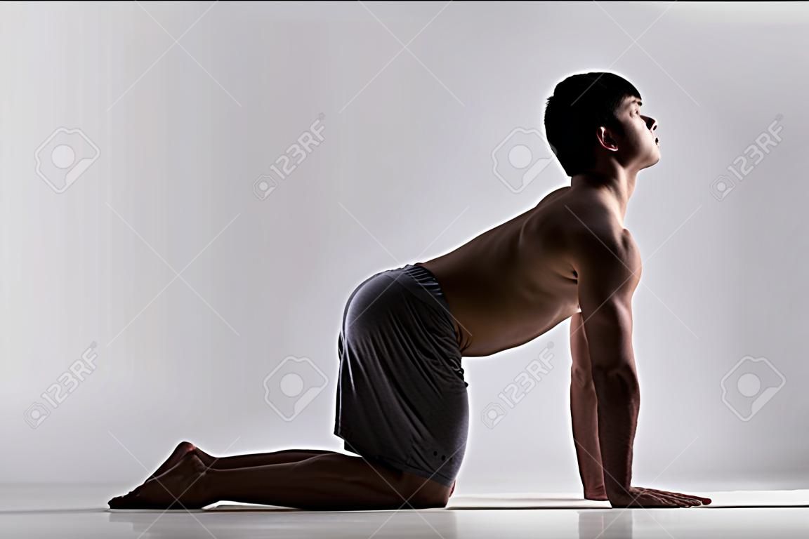Sporty muscular young man working out, yoga, pilates, fitness training, bend in Cow yoga posture, Bitilasana, asana often paired with Cat Pose on the exhale, gray background, low key shot