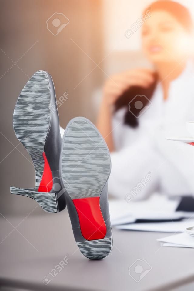 轻松的女实业家与腿坐书桌在咖啡休息期间在办公室。时髦的鞋的企业夫人有红色脚底和高跟鞋的喝茶的。休息为疲倦的女性双腿概念