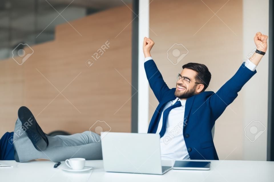 Satisfied businessman happy to finish work with laptop at office, raises hands and puts feet up on table, relaxing after hard working day in expectation of weekend leave, relaxed workday, no stress
