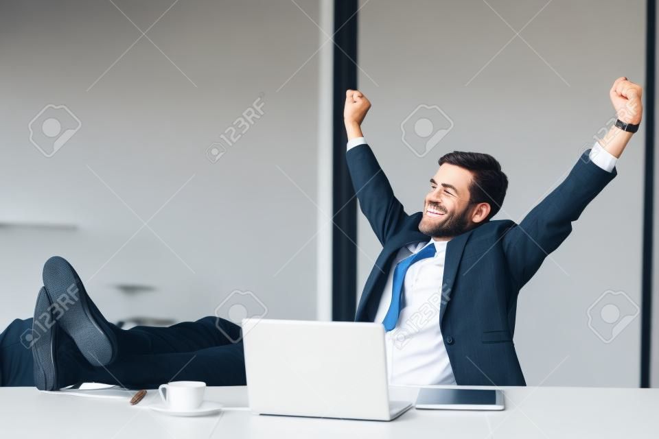 满意的商人很高兴在办公室完成与笔记本电脑的工作，举起双手，将脚放在桌子上，在辛勤工作一天后放松，期望周末休假，放松工作日，没有压力