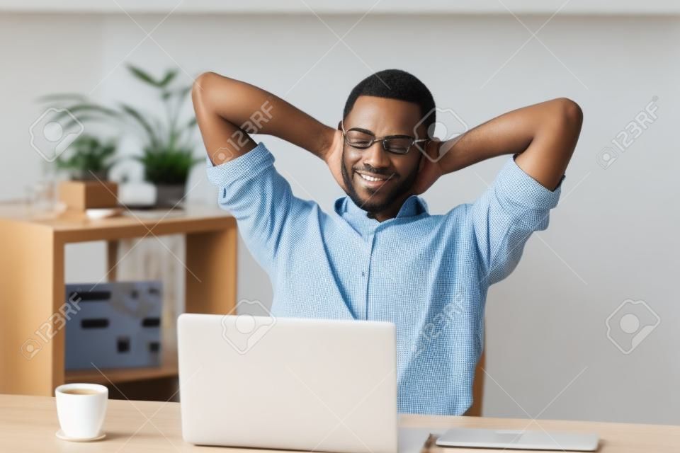 Zadowolony szczęśliwy atrakcyjny Afroamerykanin relaksujący ręce za głową przy kawie i laptopie po przerwie w domowym biurku, czarny zrelaksowany biznesmen cieszący się przyjemnym dniem w pracy