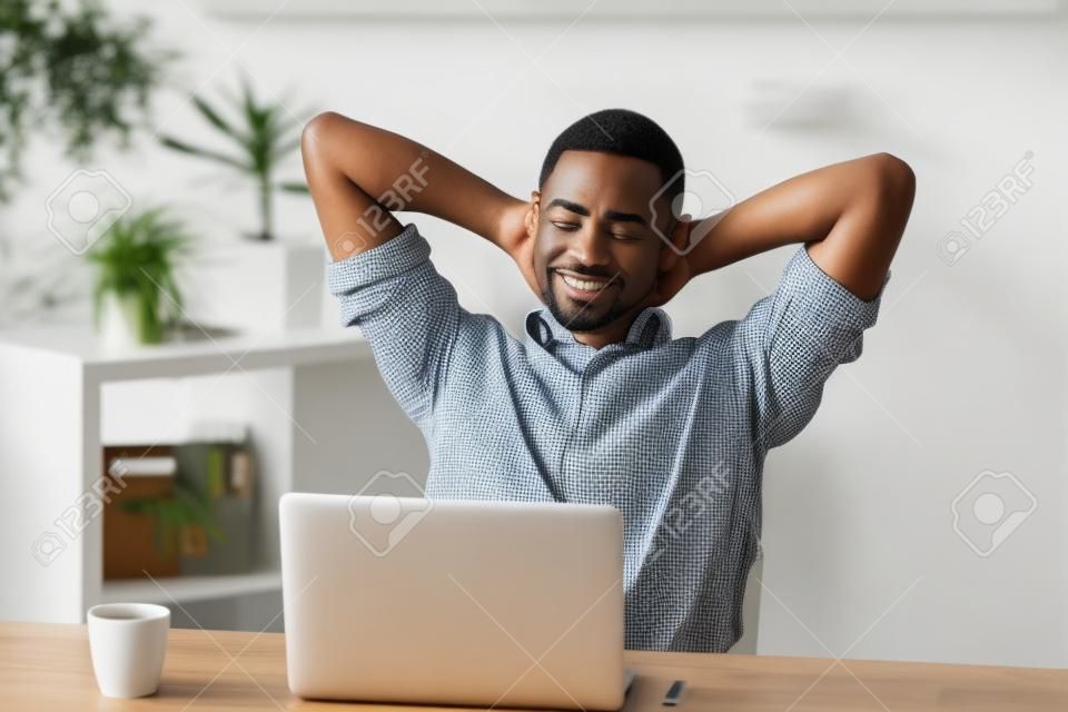 Uomo afroamericano attraente felice soddisfatto che si rilassa le mani dietro la testa con caffè e computer portatile che hanno la scrivania dell'ufficio della rottura a casa, uomo d'affari rilassato nero che gode del buongiorno piacevole sul lavoro
