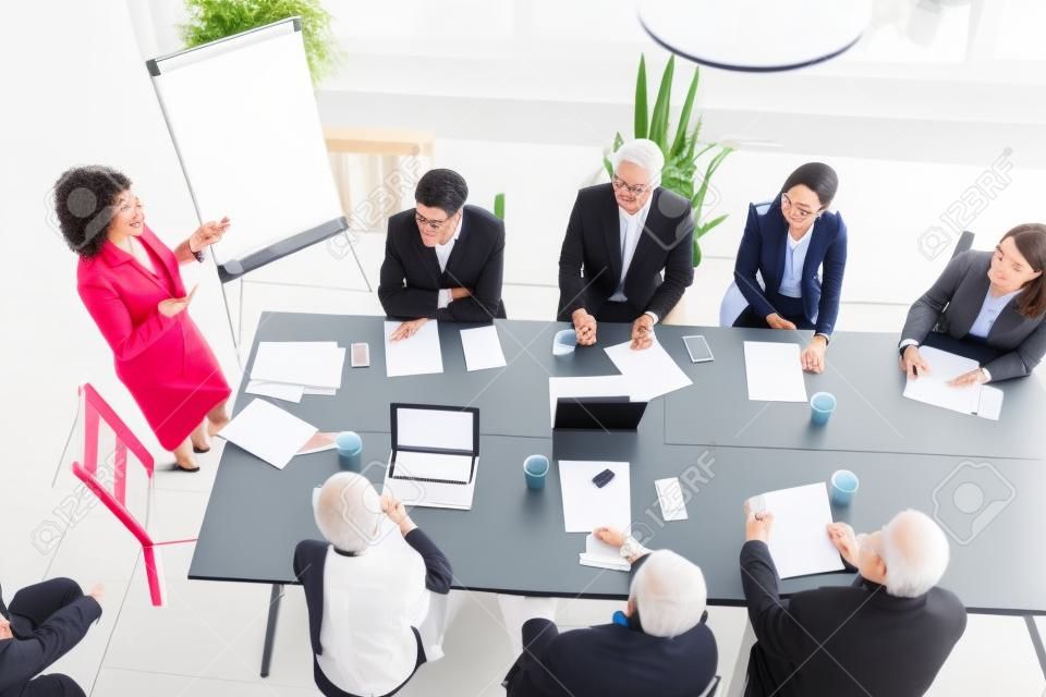 老年的高级女商人在多种族小组办公室会议，女团队负责人，公司老板或商业教练向公司高管在会议室，顶视图中提交演示文稿