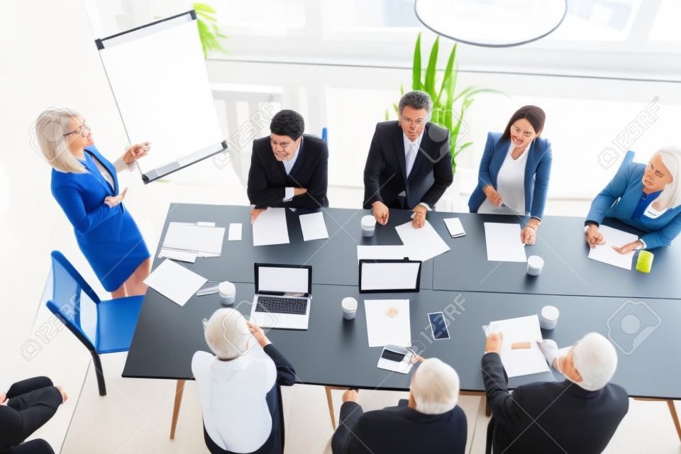 Aged senior businesswoman giving presentation at multiracial group office meeting, female team leader, company boss or business coach presenting corporate plan to executives in boardroom, top view