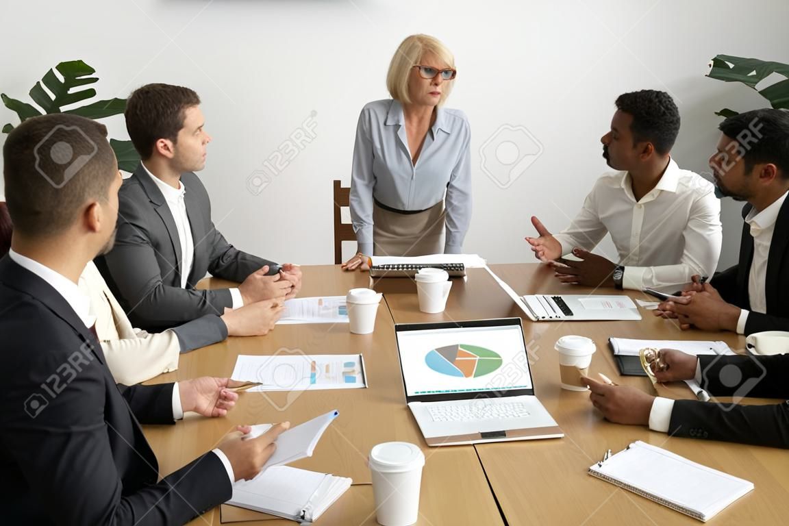 Serious aged businesswoman leading corporate team meeting talking to multiracial employees, senior female boss ceo leader discussing work with diverse subordinates at company group briefing