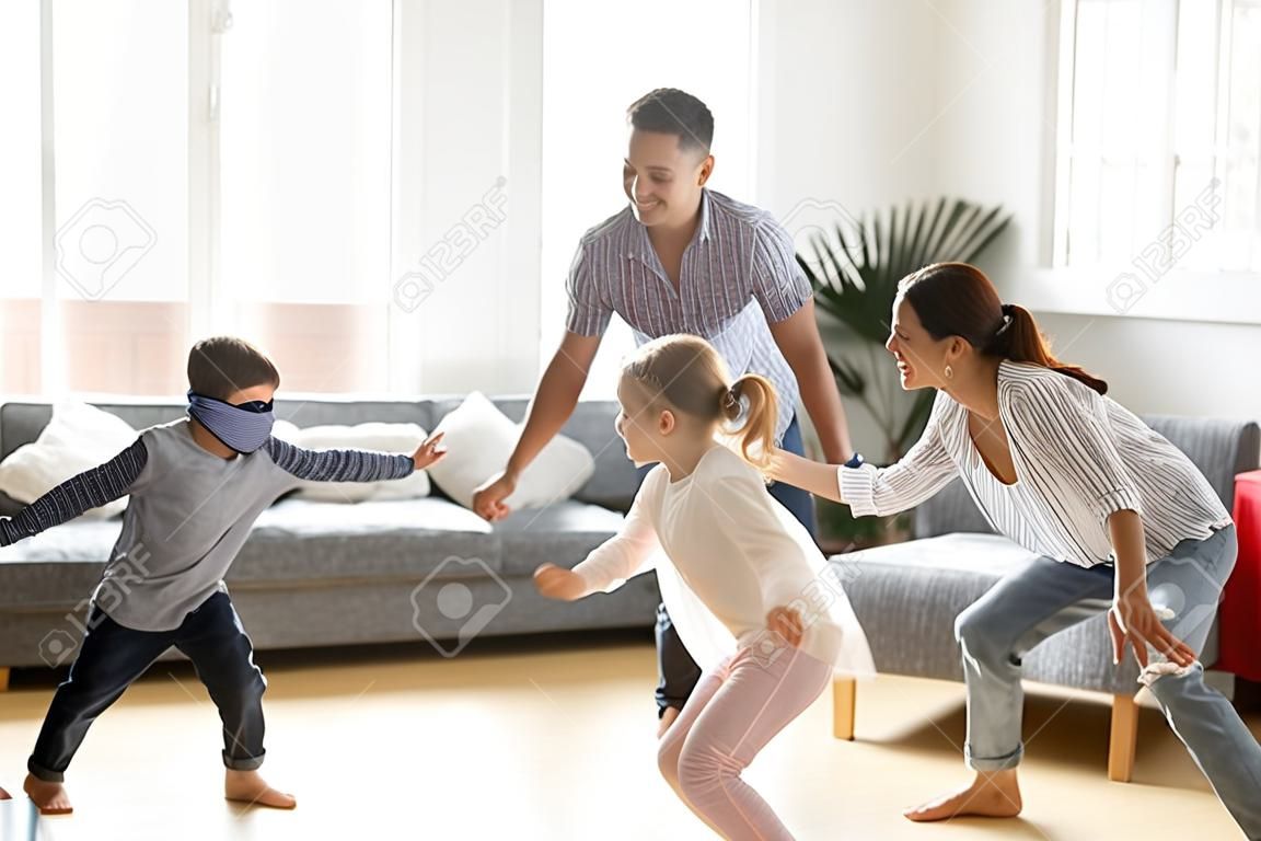 Милый маленький мальчик с завязанными глазами играет в прятки дома, родители и дети смеются, проводят время вместе, наслаждаясь игрой на выходных, счастливая семья из четырех человек веселится на досуге в гостиной